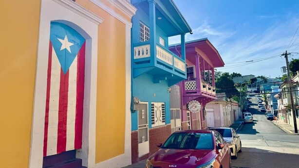 Una calle con edificios de colores, un coche rojo estacionado al lado y un cielo azul despejado.
