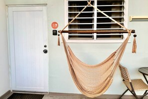 Una habitación con una puerta blanca, una ventana con persianas, un sillón hamaca y una mesita.
