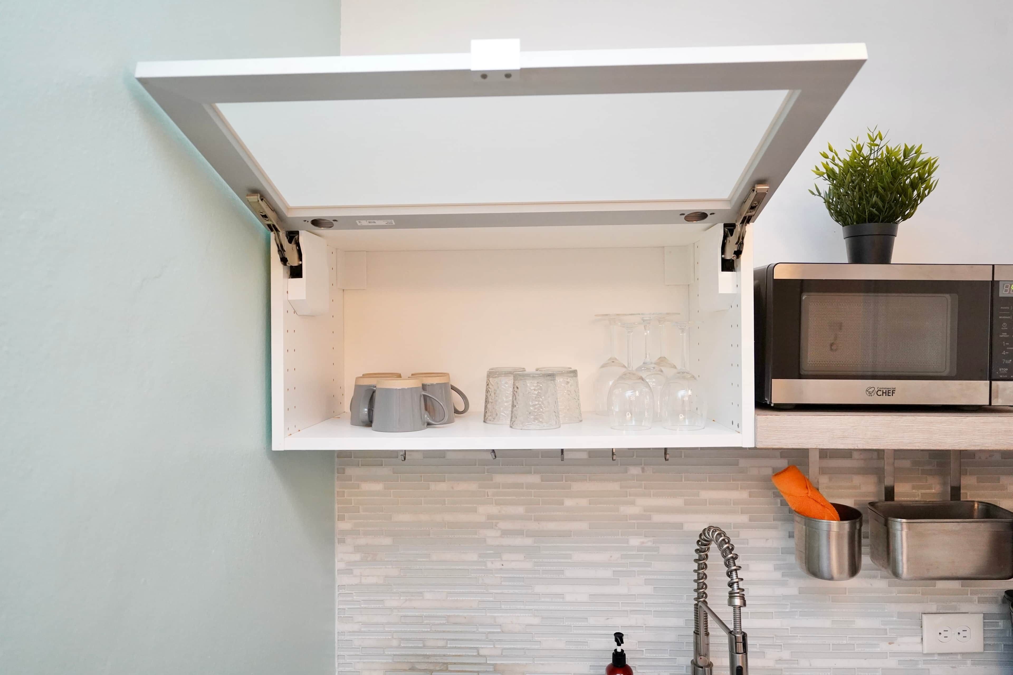 A kitchen with a built-in microwave, a hanging pot rack, and a cabinet with mugs and glassware.