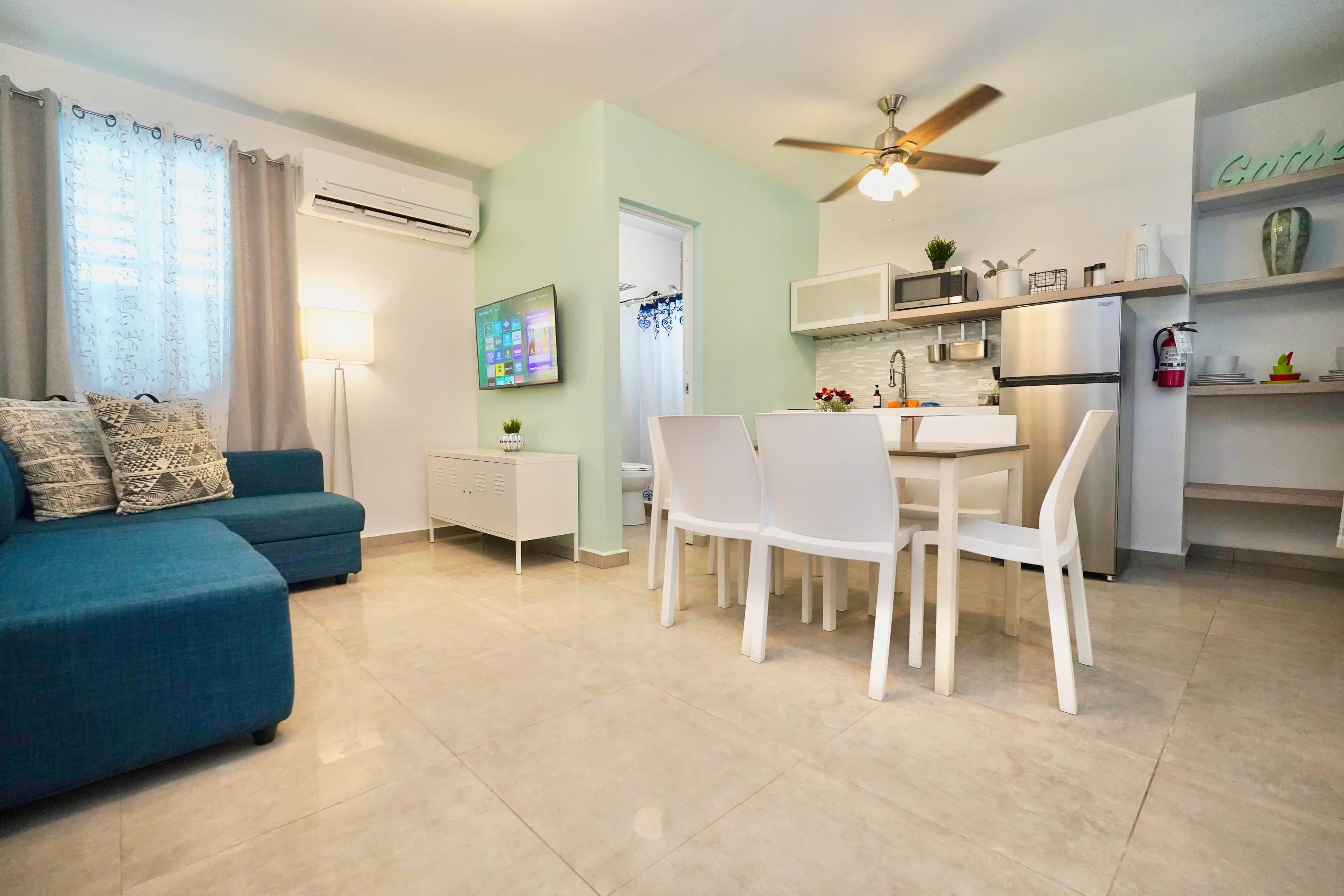 Una sala moderna con un sofá azul, un comedor con sillas blancas, una cocina con refrigerador y microondas, y un ventilador de techo.