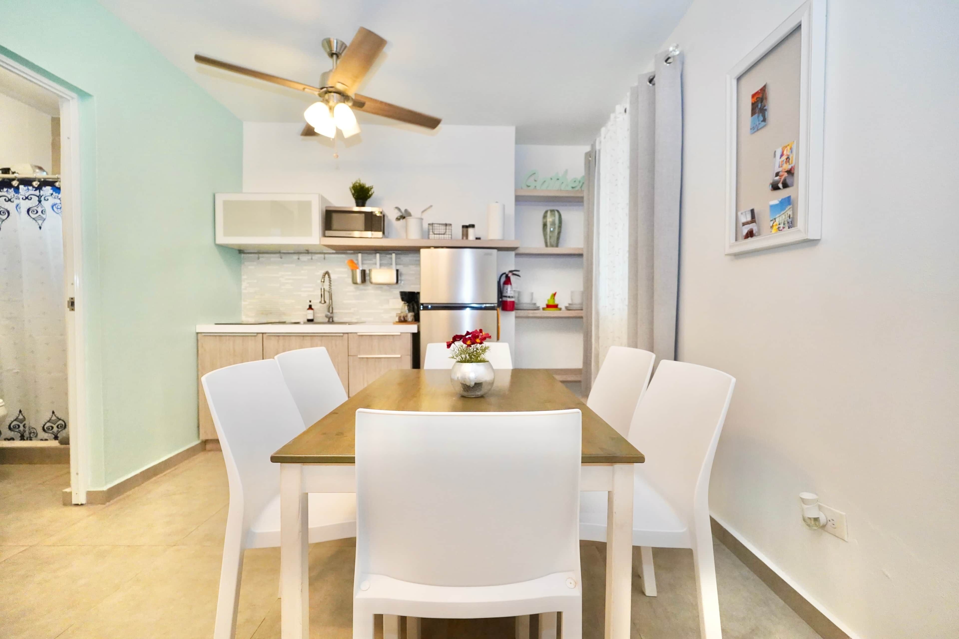 Un coin repas avec une table en bois et des chaises blanches, un ventilateur de plafond et une cuisine en arrière-plan.