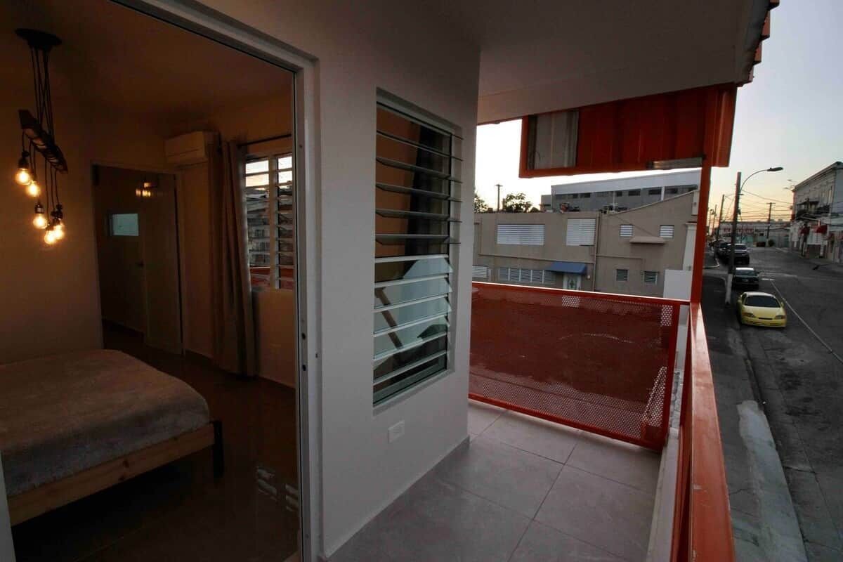 A balcony with a red safety barrier, a bed, and a window with horizontal blinds.