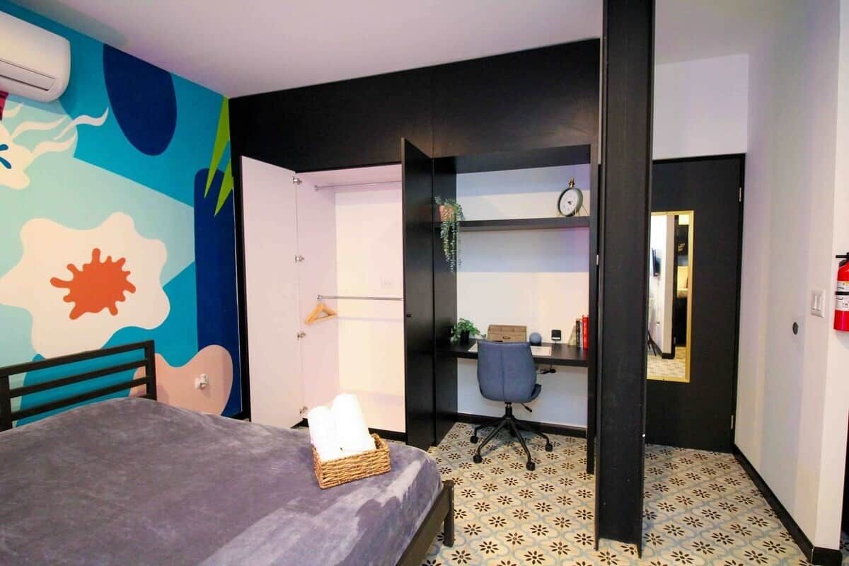 A bedroom with a bed, a desk, a chair, a wall clock, and a patterned floor.
