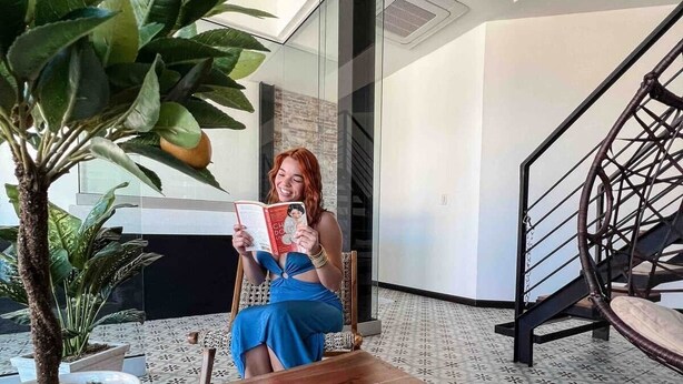 A woman reading a magazine in a modern room with a staircase and potted plants. - 5.2 Loft • Lobby • Generator • Parking • 2nd Floor (Mayagüez)