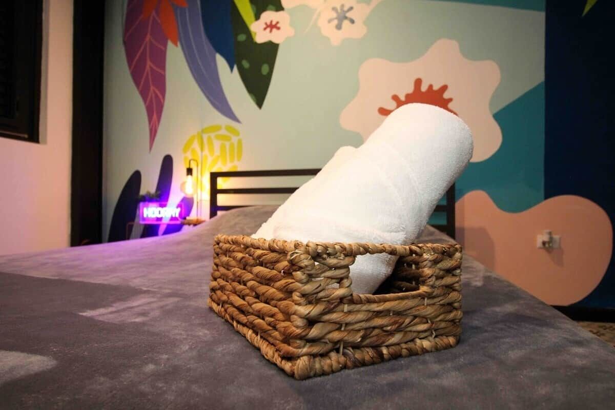 Ett rum med en flätad korg som innehåller en hoprullad vit handduk på en säng, en neonreklamskylt i bakgrunden och en vägg med en väggmålning med blommotiv.