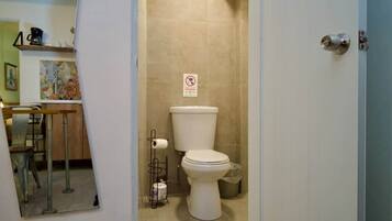 A bathroom with a toilet, a trash bin, a paper towel holder, and a no-smoking sign.