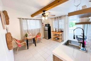 A compact kitchen with a refrigerator, a dining table with chairs, a ceiling fan, and a window with curtains.