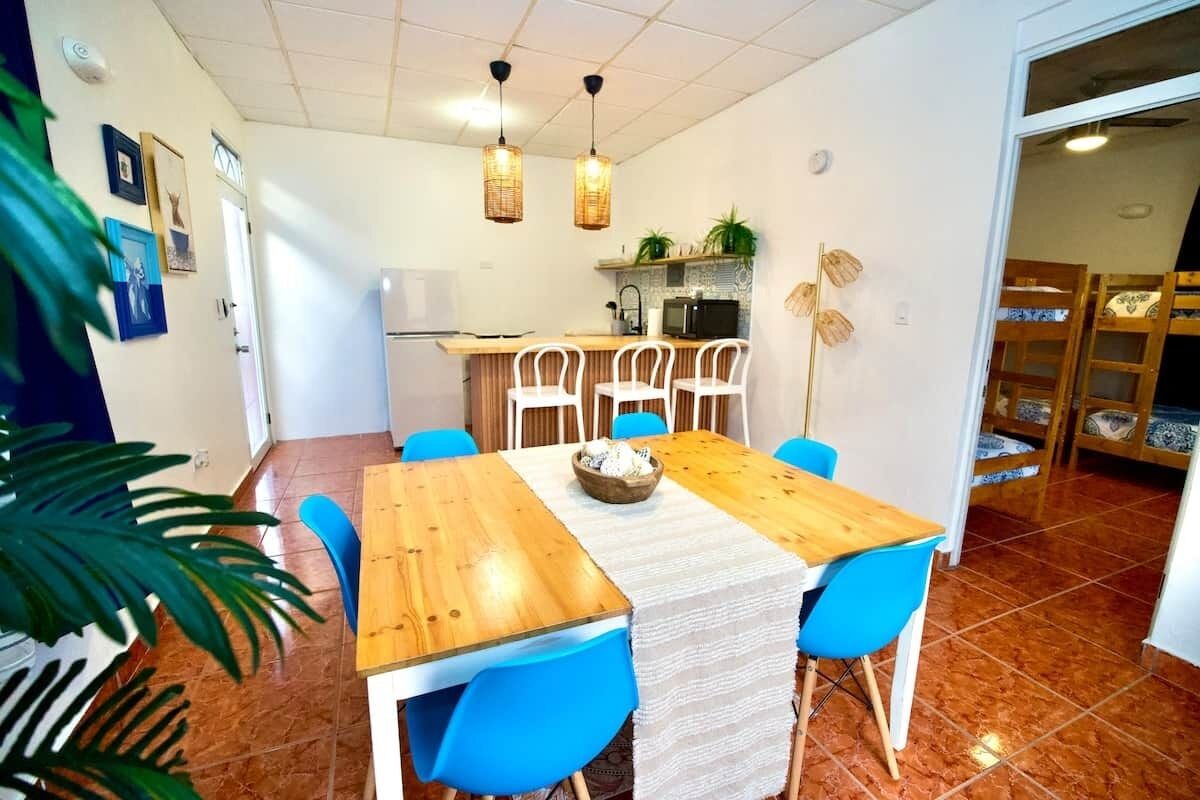 A dining area with a wooden table, blue chairs, a kitchen in the background, and a plant on the left.