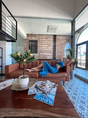Un salón moderno con un sofá de cuero marrón, una planta en maceta, revistas y un ventanal con vistas.