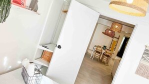 Une pièce avec une porte blanche, un panier rempli de bouteilles, une plante suspendue et un coin repas avec une table et des chaises.
