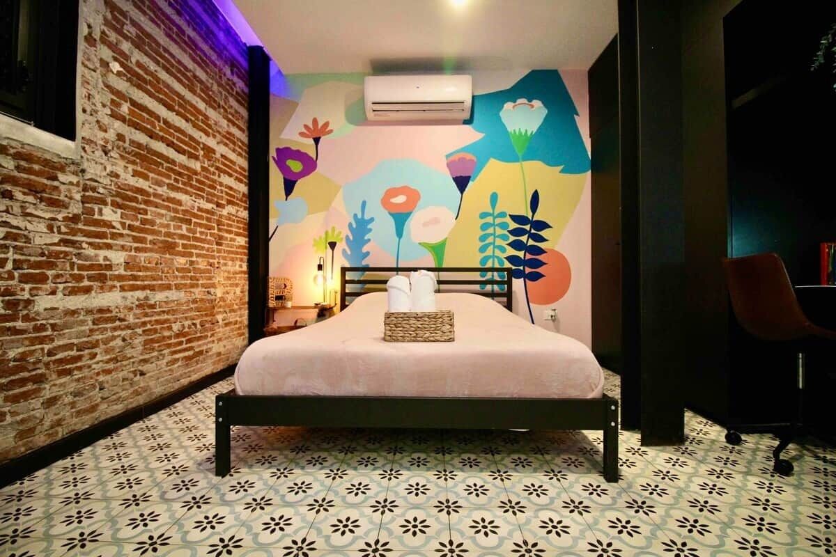 A room with a bed, a brick wall, a colorful mural, and patterned floor tiles.