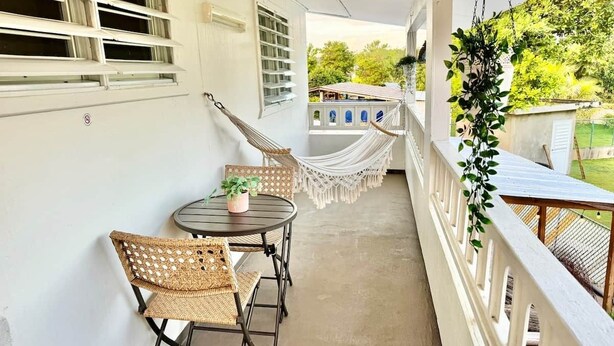 A balcony with wicker furniture, a hanging chair, and a view of a house and greenery. - 3.5 Near Beach • Generator • Hammock • 2nd floor (Miradero)