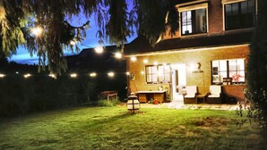 A house with a brick exterior, a porch with chairs, and a well-lit yard at dusk.