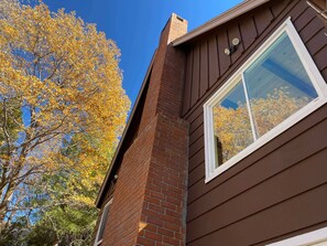 A corner of a house with a brick chimney, a window with white trim, and a tree with yellow leaves. - Lake Arrowhead Mountain Hideaway by Navigate Stays (Lake Arrowhead)