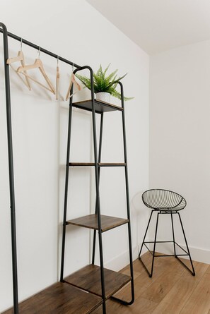 Una estantería metálica negra con perchas de madera y una planta en maceta encima, un taburete de alambre negro y suelo de madera.