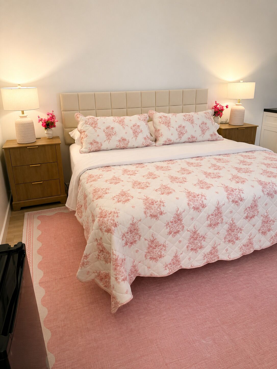 A bedroom with a bed, two bedside tables, a nightstand, a pink rug, and a wall-mounted lamp.
