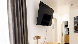 Un salon moderne avec un bureau en bois, un téléviseur à écran plat fixé au mur, un fauteuil en cuir et une grande fenêtre avec des rideaux.