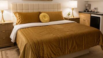 A hotel room with a large bed, a cityscape painting, a small kitchenette, and a bedside table with a lamp.