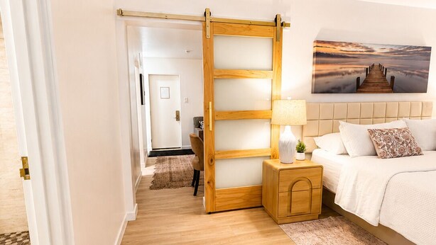 Une chambre avec un plancher en bois, un lit recouvert de linge de lit blanc, une table de chevet avec une lampe et un tableau représentant une jetée accroché au mur.