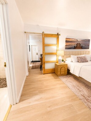 A bedroom with a wooden floor, a bed with white bedding, a nightstand with a lamp, and a painting of a pier on the wall. - Tex-Haus (Snyder)