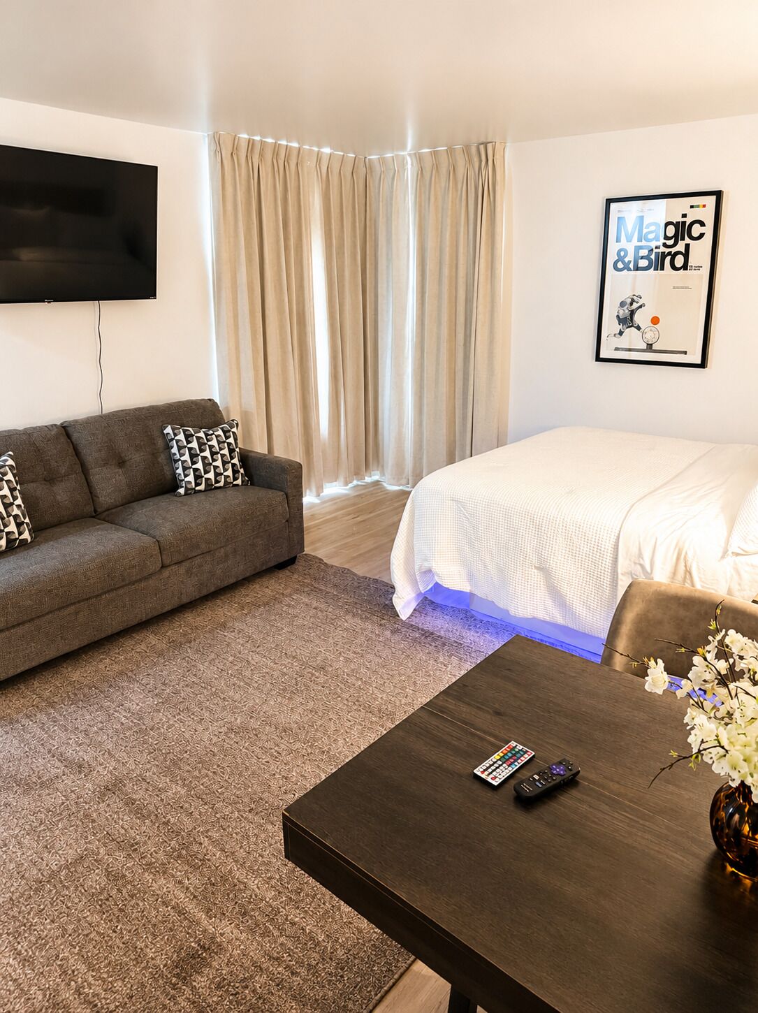 Ein Hotelzimmer mit einer Couch, einem Bett, einem an der Wand montierten Fernseher, einem gerahmten Bild und einem Tisch mit einer Blumenvase.