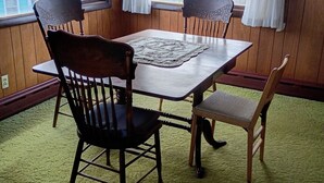 Uma área de refeições com mesa de madeira, quatro cadeiras, um lustre estampado e grandes janelas com cortinas.