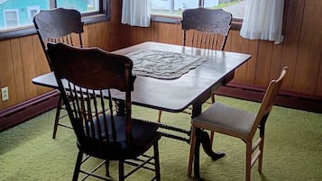 A dining area with a wooden table, four chairs, a patterned chandelier, and large windows with curtains.