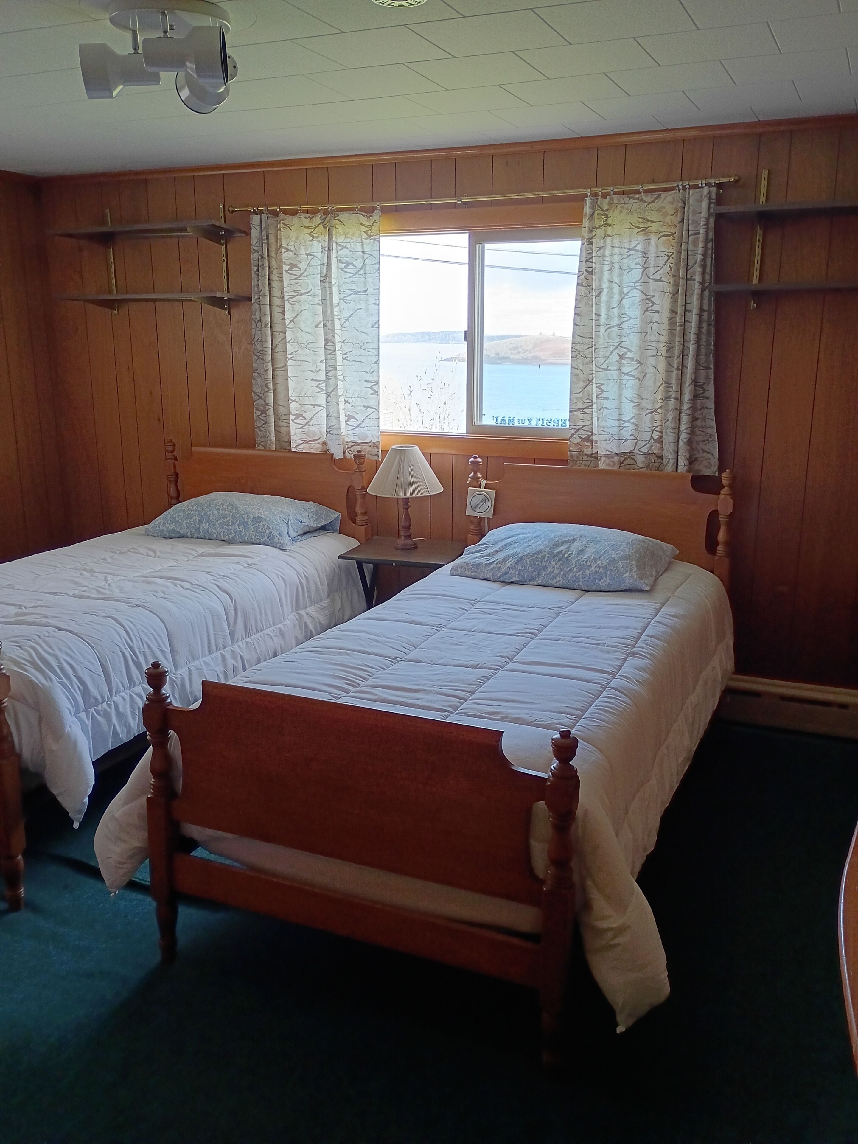 A room with two beds, wooden headboards, a nightstand with a lamp, and a window with curtains.