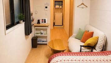 A compact hotel room with a bed, a small table, a desk with a computer, a clock, and a window with curtains.