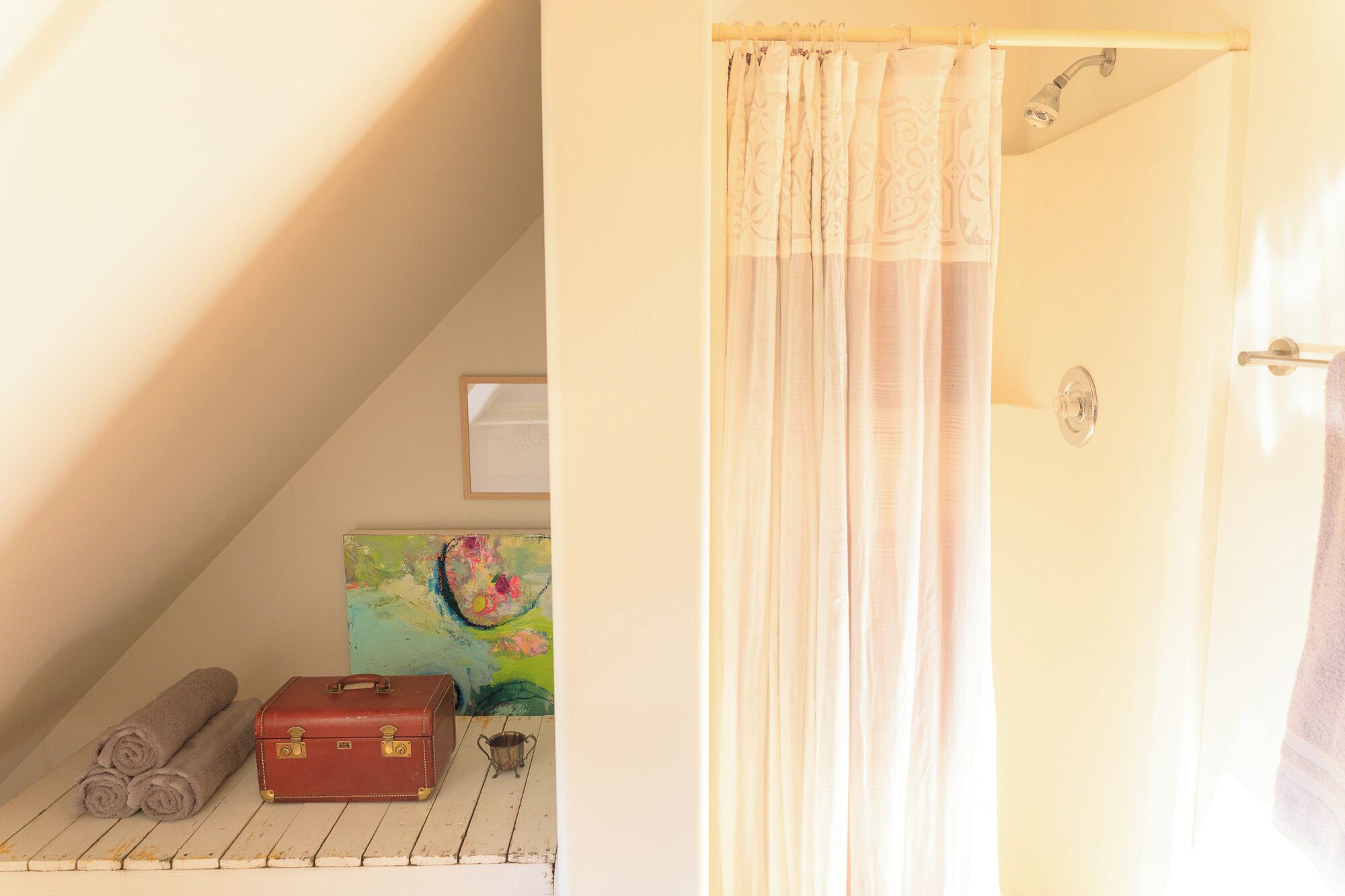 Um quarto com cortina de chuveiro, uma mala vermelha, um balde de metal e um quadro colorido numa prateleira de madeira.