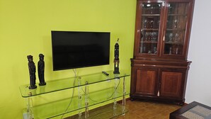 Een woonkamer met een flatscreen televisie aan een groene muur, een glazen displaykast met beeldjes, en een houten kast met glazen deuren.