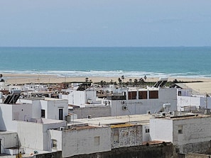 Uma área residencial costeira com edifícios brancos, alguns com telhados planos e outros com telhados inclinados, com vista para uma praia e o oceano ao fundo.