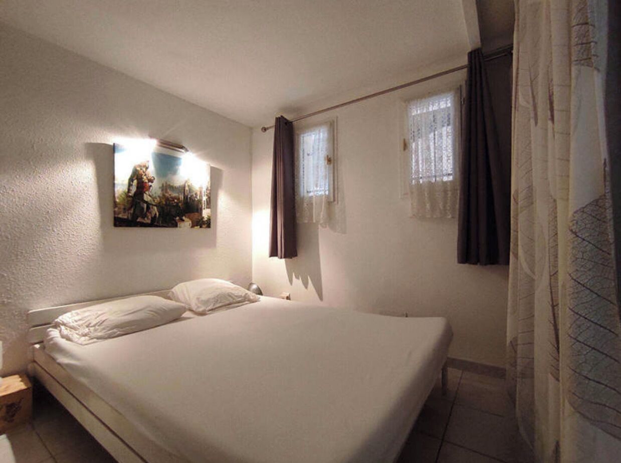 Une chambre avec un lit simple, une table de chevet, une lampe fixée au mur et une fenêtre avec des rideaux.