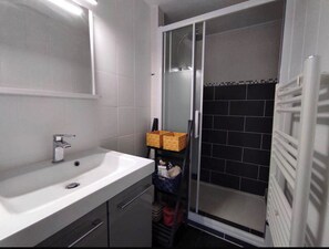 Casa de banho com lavatório branco, espelho, chuveiro com porta de vidro e porta-toalhas.