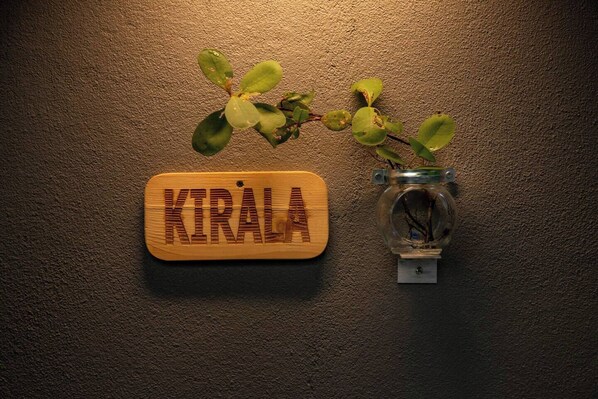 Un rètol de fusta amb la paraula 'KIRALA' fixat a una paret rugosa, al costat d'un bocal amb una planta.