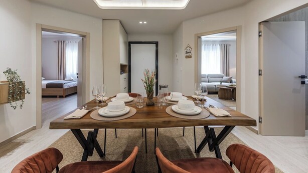 A dining area with a wooden table set for four, surrounded by chairs, with a view into adjacent rooms. - Loen Modern Seaside 3BR Apartment by PikHost (Durrës)