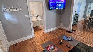Ett vardagsrum med en platt-TV, ett soffbord med tidningar och en hall som leder till ett sovrum.