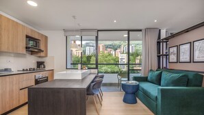 A modern kitchen with a built-in microwave, a dining area with a green sofa and chairs, and a large window offering a city view. - GRAND BY LINARES  (Medellín)