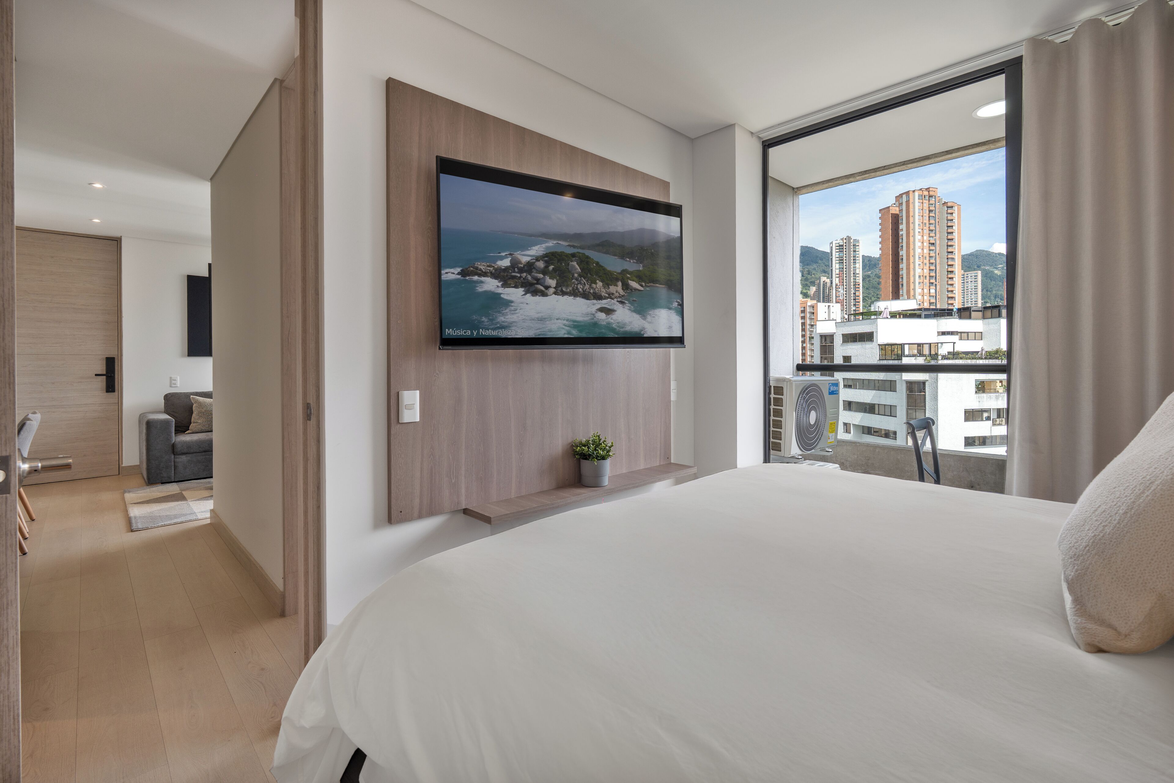 Una habitación de hotel moderna con una cama grande, un televisor montado, un balcón con vista a la ciudad y una pequeña planta en maceta.