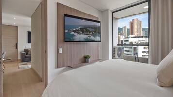 Una habitación de hotel moderna con una cama grande, un televisor montado, un balcón con vista a la ciudad y una pequeña planta en maceta.