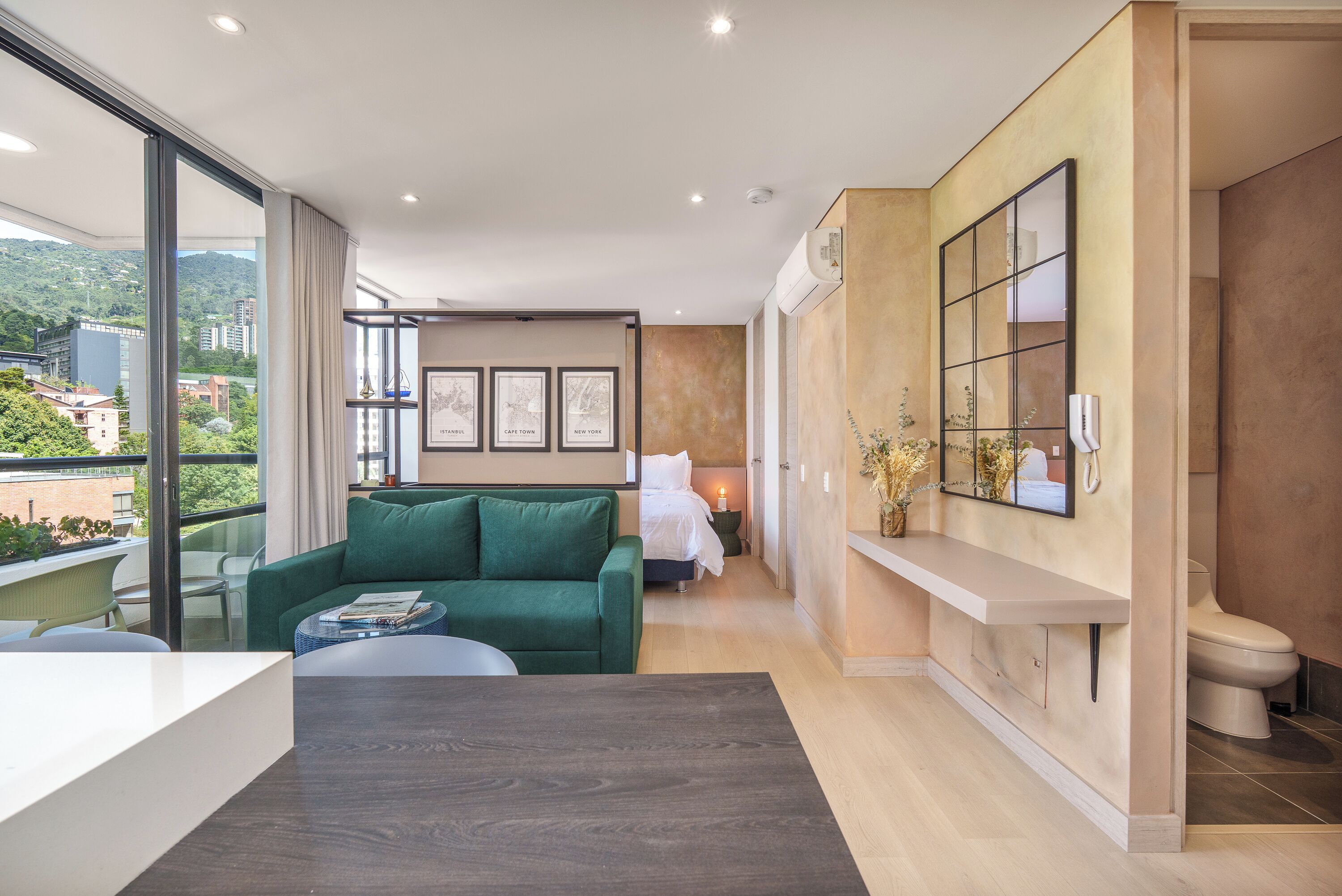 Een moderne hotelkamer met een groene bank, een klein tafeltje, een bed met witte lakens, een groot raam en een badkamer met toilet en wastafel.