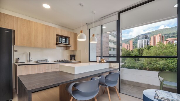 A modern kitchen with a central island, wooden cabinets, and a view of a cityscape through large windows. - GRAND BY LINARES  (Medellín)