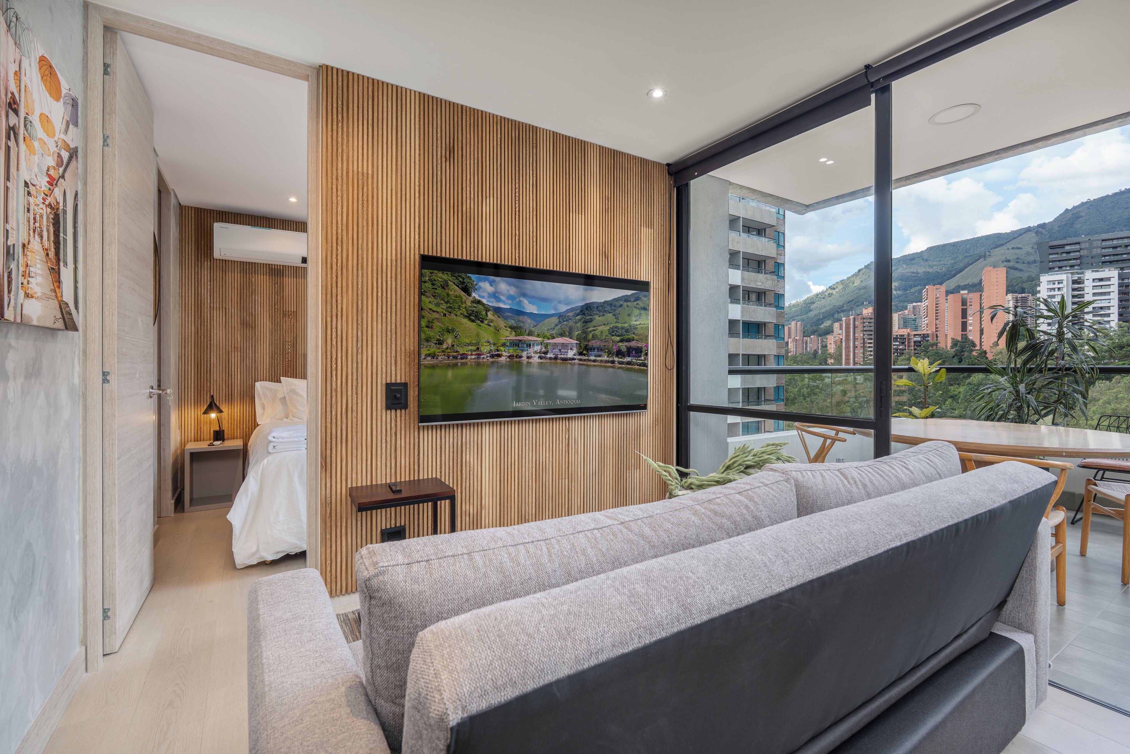 A modern hotel room with a large TV mounted on a wooden panel wall, a sofa, a small table, and a view of a cityscape and mountains.