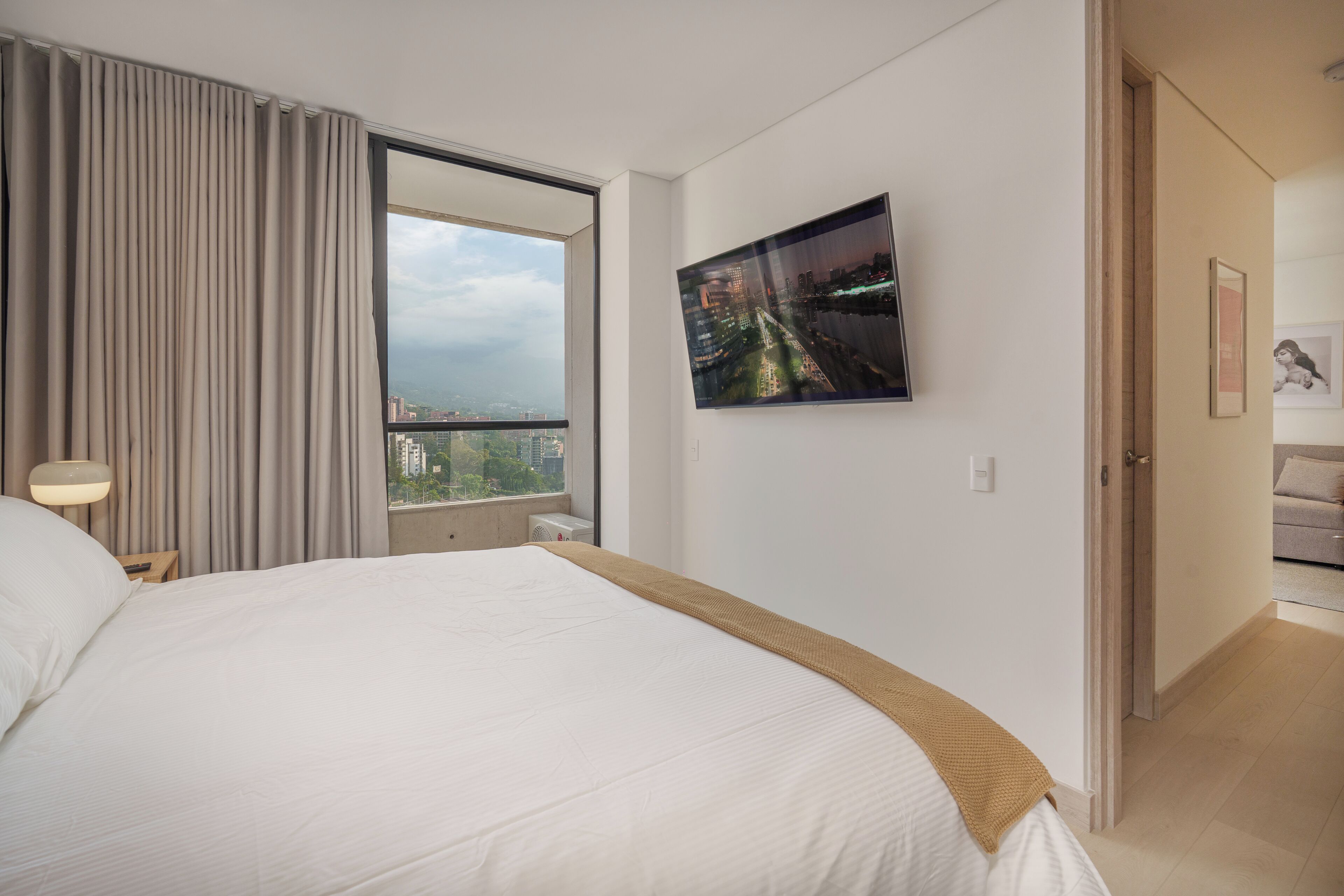 Ein Hotelzimmer mit einem großen Bett, einem Fenster mit Vorhängen, einem an der Wand montierten Fernseher und einer Tür zu einem weiteren Raum.