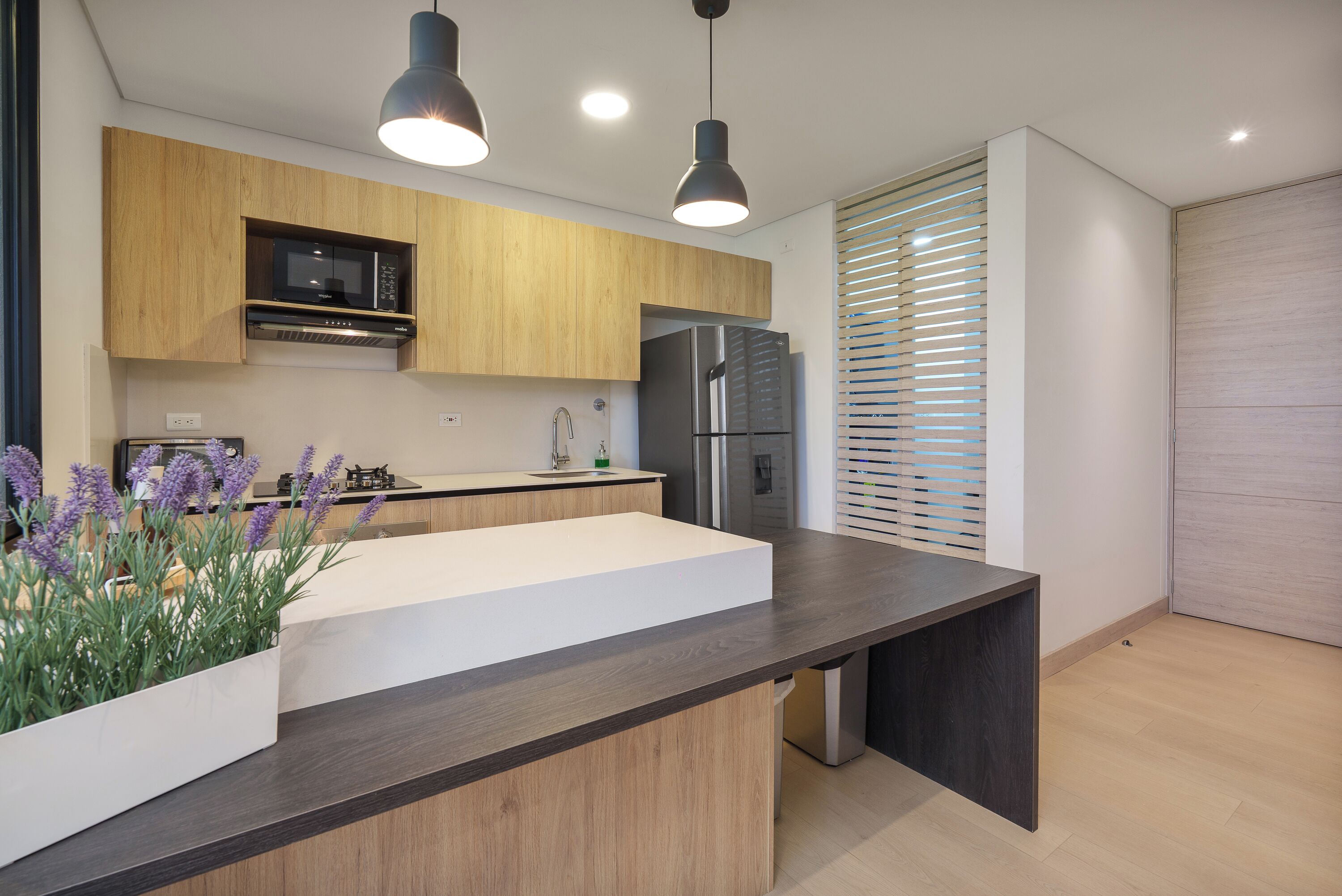A modern kitchen with a central island, wooden cabinets, a built-in microwave, and a stainless steel refrigerator.