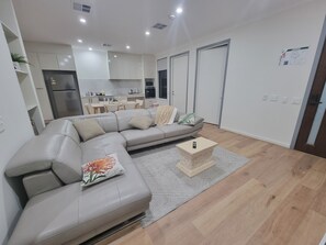 Een moderne woonkamer met een grijze bank, een klein houten salontafeltje en een keuken op de achtergrond.