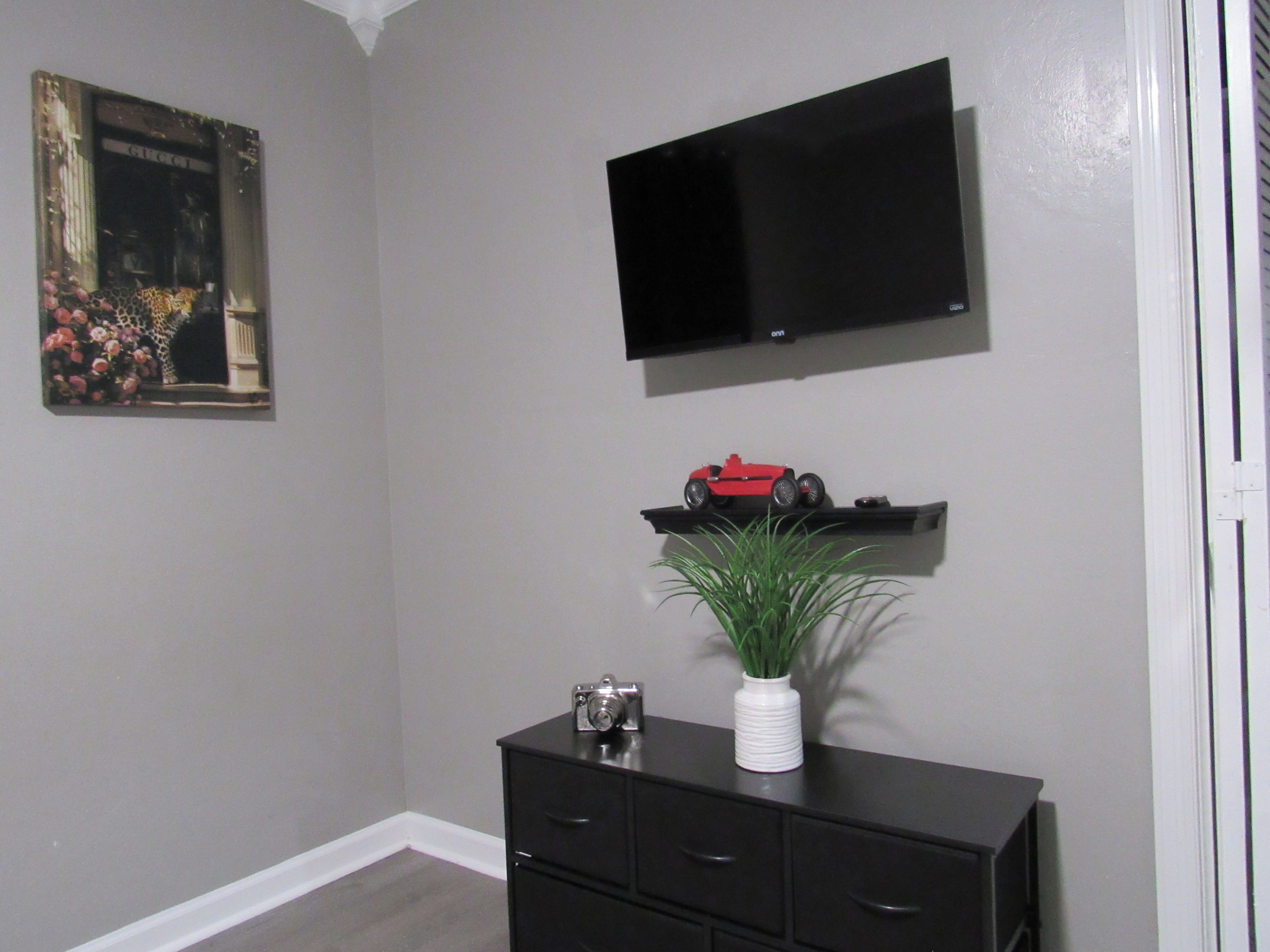 A room with a black TV on the wall, a small red toy car on a shelf, a white vase with a plant, and a black dresser with drawers.