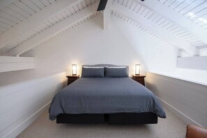 Een slaapkamer met een schuine kap, een bed met grijze beddengoed, twee bedlampen en een bruine leren fauteuil.
