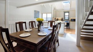 Un comedor con una mesa de madera dispuesta para cuatro, un florero con flores amarillas y una cocina al fondo.