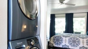 Habitación con lavadora, cama con ropa de cama estampada, ventilador de techo y ventana con cortinas.